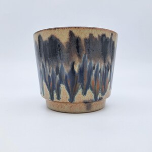 Op de afbeelding: Een keramieken beker met een gespikkelde beige basis en een dramatisch, donkerblauw en zwart glazuur dat vanaf de rand naar beneden druipt. De beker heeft een brede opening en een opvallende, getrapte voet met een reactieve, meerkleurige afwerking.