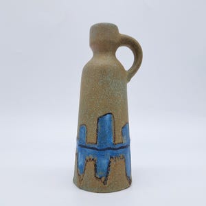 May include: A tan ceramic vase with a handle. The vase has a cylindrical shape with a flared base. It features a blue and brown abstract design around the base. The vase is approximately 8 inches tall.