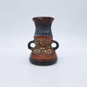 Puede incluir: Un jarrón de cerámica en tonos marrón, negro y beige. El jarrón tiene una parte superior acampanada, dos asas pequeñas y una superficie texturizada. La base es negra, con una sección media marrón y una parte superior marrón oscuro.