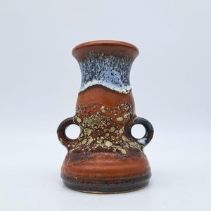 Puede incluir: Un pequeño jarrón de cerámica marrón con dos asas y un esmalte azul y blanco. El jarrón tiene una textura moteada.