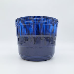 May include: A blue ceramic plant pot with a glossy finish. The top section has a darker, dripped glaze effect, fading to a lighter blue at the base. Horizontal ridges are visible around the top and bottom.