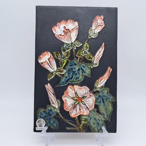 May include: A rectangular black ceramic wall plaque featuring a raised, hand-painted floral design of pink and white morning glory flowers with green leaves. A small circular sticker with the text "RUSC ART" is visible in the bottom left corner.