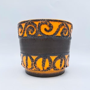 Può includere: Fioriera cilindrica in ceramica con una fascia marrone scuro e dettagli a motivi arancioni e neri. La fioriera ha una superficie strutturata e un'ampia apertura, adatta a varie piante. Il design presenta un motivo a spirale ripetuto.