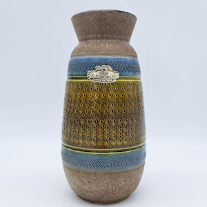Op de afbeelding: Een bruine keramische vaas met een geel en blauw geometrisch patroon. De vaas heeft een label met de tekst "Bay Keramik Handgemacht".