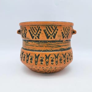 May include: A terracotta planter with black geometric designs and two small handles. The planter has a textured surface and a slightly rounded shape.
