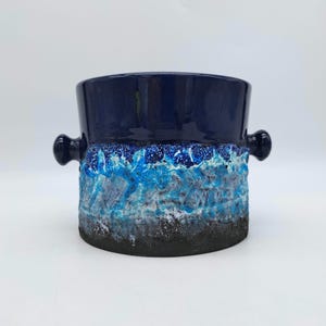 Puede incluir: Un jarrón de cerámica con una parte superior azul oscuro y dos asas pequeñas. El cuerpo del jarrón presenta un diseño texturizado con tonos de azul, blanco y negro, creando un efecto en capas. El jarrón tiene forma redonda.