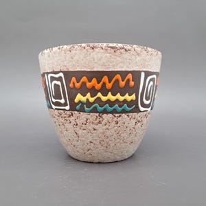 Può includere: Una tazza in ceramica con una finitura testurizzata maculata beige e marrone. Una larga fascia marrone scuro circonda il centro, caratterizzata da linee ondulate in rilievo color arancione, giallo e turchese, affiancate da motivi a spirale bianchi.