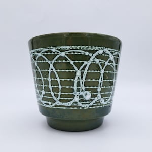 Pode incluir: Um vaso de cerâmica verde escuro com um design de linhas pontilhadas brancas interligadas na parte superior. O vaso tem uma borda ligeiramente alargada e uma base de pedestal. O fundo é branco liso.