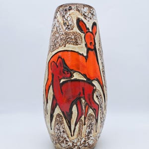 Puede incluir: Un jarrón de cerámica alto con un exterior beige y marrón moteado y texturizado. El jarrón presenta una ilustración estilizada de dos ciervos en tonos rojos y naranjas. Los ciervos están delineados en negro.