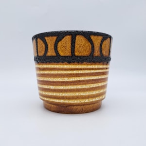 Peut inclure: Un cache-pot cylindrique en céramique doté d'un bord noir texturé orné d'un motif en arc répété. Le corps du pot présente une série de rayures horizontales blanches superposées sur une finition émaillée brun moyen avec une texture mouchetée.