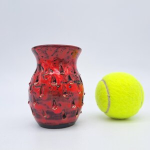May include: A small, red ceramic vase with a textured surface and a yellow tennis ball. The vase has a dark base and a wide opening. The tennis ball is a bright yellow colour.