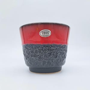 Peut inclure: Un cache-pot en céramique avec une partie supérieure rouge lisse et brillante et une partie inférieure gris foncé texturée. Un autocollant ovale argenté avec le texte noir Jopeko Keramik est centré sur la section rouge du récipient.