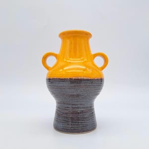 Puede incluir: Un jarrón de cerámica con dos asas, con una parte superior anaranjada ancha y un cuerpo marrón oscuro texturizado. El jarrón tiene forma cilíndrica con una parte superior ensanchada y dos asas circulares. La parte inferior del jarrón es de color marrón oscuro.