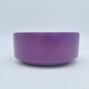 May include: A round, deep bowl with a smooth, solid purple finish. The bowl features straight, vertical sides that curve slightly inwards at the base, creating a clean, uniform silhouette. The surface has a subtle, matte-like texture.
