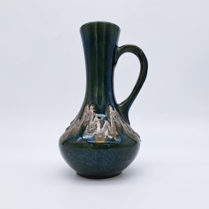 Puede incluir: Jarra de cerámica verde oscuro con asa. La jarra tiene una base bulbosa y un cuello estrecho. El borde y el asa son de color verde oscuro, mientras que la base presenta un diseño texturizado en marrón claro y dorado.