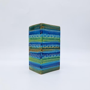May include: A rectangular ceramic vase with a blue, green, and brown color scheme. The vase features horizontal bands of varying shades of blue and green, adorned with repeating geometric patterns. The vase has a square base and a slightly textured surface.