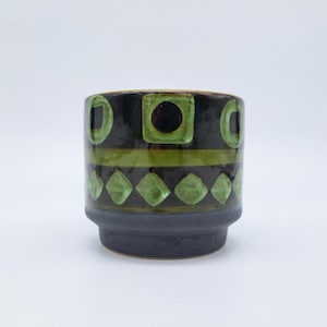 Può includere: Una tazza cilindrica in ceramica con base grigio scuro e sezione superiore nera. La tazza presenta motivi geometrici verdi, tra cui quadrati, cerchi e diamanti, e una striscia verde orizzontale. Il bordo è di colore marrone chiaro.