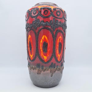 May include: A tall, cylindrical ceramic vase with a vibrant red glaze. The vase features a repeating pattern of black, textured, oval shapes with orange and yellow centres. The base is a speckled grey, and the top has a dark rim.