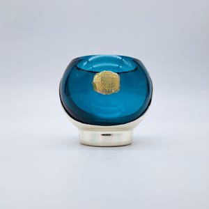 Può includere: Una ciotola di vetro color turchese con base argentata. La ciotola ha una forma arrotondata e una piccola etichetta dorata è attaccata sul davanti. L'oggetto è probabilmente un oggetto decorativo o un piccolo contenitore.