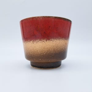 Può includere: Un vaso in ceramica dalla forma svasata, con una sfumatura di colori. La parte superiore è rosso intenso, che sfuma in un centro beige e termina con una base marrone scuro. Il vaso ha una superficie strutturata.