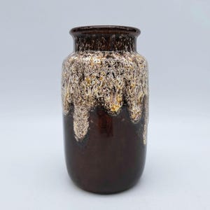 Puede incluir: Un jarrón de cerámica marrón con un esmalte blanco y amarillo. El esmalte tiene un efecto de goteo, creando un aspecto único y rústico.