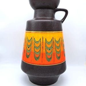 May include: A brown ceramic vase with a handle and a wide base. The vase features a vibrant orange and yellow band with a repeating green design. The vase is marked "XXL" at the bottom.