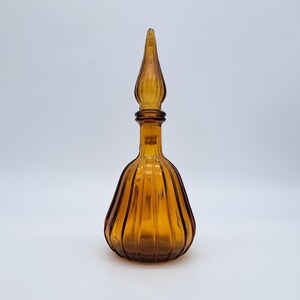 May include: Amber-colored glass perfume bottle with a ribbed body and a tall, pointed stopper. The bottle has a bulbous shape and a long, slender neck. The stopper is also amber-colored and has a decorative, pointed top. The bottle is likely vintage.