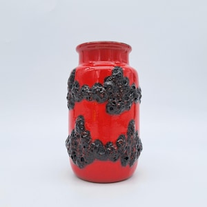 Puede incluir: Un jarrón de cerámica rojo vibrante con un acabado brillante. El jarrón presenta un diseño texturizado, gris oscuro, similar a la lava alrededor del cuerpo. El jarrón tiene forma cilíndrica con un borde ligeramente ensanchado, ideal para flores o como pieza decorativa.