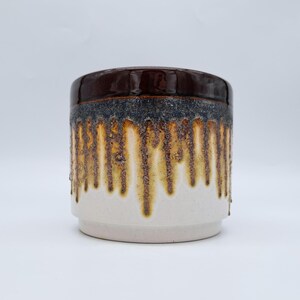 May include: A ceramic jar with a dark brown lid and a white base. The middle section has a textured design with brown and yellow drips over a dark grey band. The jar is cylindrical in shape.