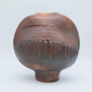 May include: A handcrafted, round ceramic vase with a textured, dark brown surface. The vase features a unique design with a lighter brown top and dark brown drips along the middle, and a small base.