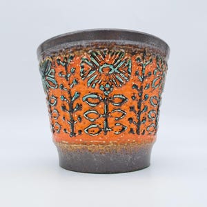 May include: A ceramic planter featuring a vibrant orange textured body with embossed floral patterns highlighted in turquoise and black. The rim and base are finished in a dark brown glaze, creating a contrast with the bright central band.