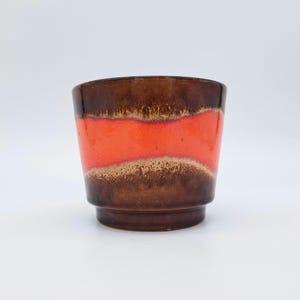 Peut inclure: Une tasse en céramique présentant une large bande horizontale de glaçure orange vif au centre, contrastée par une glaçure tachetée brun foncé sur les sections supérieure et inférieure. La tasse a une forme cylindrique avec une base légèrement effilée.
