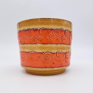 May include: A cylindrical ceramic planter featuring a vibrant orange and tan glaze. The exterior is decorated with two horizontal bands of embossed, overlapping circular patterns that wrap around the body of the vessel, creating a textured relief.