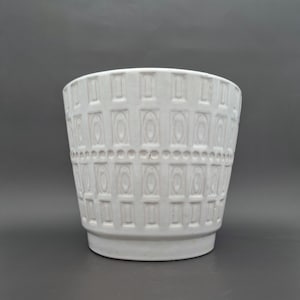 Peut inclure: Un pot de fleurs en céramique blanche avec un motif géométrique texturé. Le pot a une forme cylindrique avec un bord légèrement évasé et une base surélevée. Le motif est composé de formes rectangulaires et circulaires.