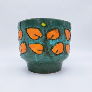 Op de afbeelding: Een keramische plantenbak met een donkergroene basis en een turquoise interieur. De buitenkant heeft oranje bladmotieven met zwarte contouren. Bovenop zit een kleine gele stip. De plantenbak heeft een licht uitlopende basis.