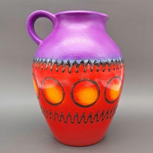 Puede incluir: Una jarra de cerámica con asa, que presenta una combinación de colores vibrantes. La parte superior es de color púrpura liso, que pasa a una base roja adornada con círculos naranjas y patrones en zigzag negros. La jarra es un artículo decorativo.