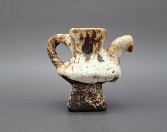 Może przedstawiać: Mały, ręcznie robiony ceramiczny imbryk o unikalnym, teksturowanym wyglądzie. Imbryk ma biało-brązową kolorystykę, z zaokrąglonym korpusem, uchwytem i dzióbkiem. Podstawa jest w kolorze ciemnobrązowym.