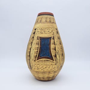 Könnte beinhalten: Eine hohe, dekorative Vase mit einer strukturierten, gelbbraunen Außenseite. Die Vase hat ein rechteckiges blaues und braunes Design in der Mitte. Der obere Rand ist rot und schwarz. Die Vase hat ein einzigartiges, handgefertigtes Aussehen.