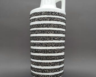 Può includere: Vaso in ceramica bianca con manico, caratterizzato da fasce orizzontali a righe bianche e nere strutturate. Il vaso ha un corpo cilindrico e una base svasata, adatto per composizioni floreali o decorazioni.