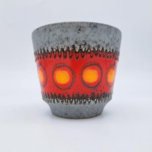 May include: A ceramic cup featuring a textured grey speckled glaze on the top and bottom sections. The centre band is a vibrant red with a row of repeating orange circular motifs, bordered by dark, jagged, tooth-like patterns above and below.