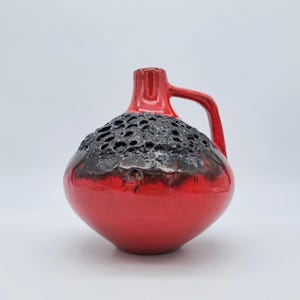 Peut inclure: Un vase en céramique rouge avec un dessus noir texturé et une anse. Le vase a une forme bulbeuse et une finition brillante. La poignée est incurvée et fixée au col du vase.