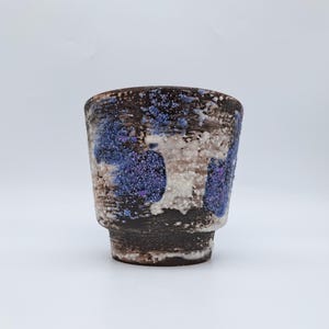 Może przedstawiać: Ręcznie wykonany ceramiczny kubek o unikalnej, teksturowanej powierzchni. Kubek ma ciemnobrązową podstawę z niebieskimi i białymi nakrapianymi akcentami. Kubek ma cylindryczny kształt z lekko rozszerzonym brzegiem, do napojów.