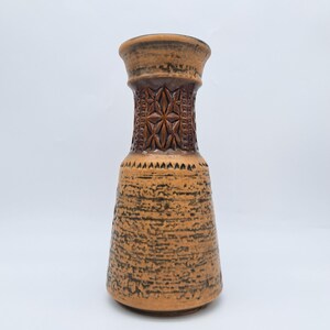 Op de afbeelding: Een hoge, bruine keramische vaas met een getextureerd oppervlak en een decoratief, gesneden bloemmotief rond de hals. De vaas heeft een uitlopende basis en een iets bredere bovenkant, met een donkerbruine rand. Het algehele ontwerp is rustiek en vintage.