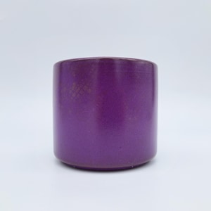 May include: A cylindrical, purple ceramic container with a slightly textured surface. The container has a rounded base and a smooth, unadorned exterior. The color is a deep, saturated purple, and the object is set against a white background.