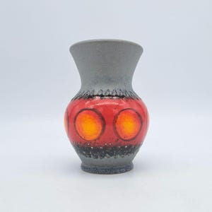 Puede incluir: Un jarrón de cerámica con la parte superior e inferior grises, una sección media roja y diseños circulares amarillos y naranjas. El jarrón tiene una superficie texturizada y una parte superior acampanada, adecuado para exhibir flores o como pieza decorativa.