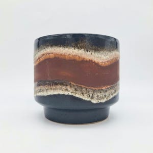 May include: A cylindrical ceramic planter with a dark blue base and rim. The body features a band of brown and beige horizontal stripes. The planter has a mid-century modern aesthetic.