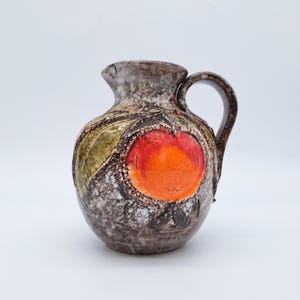 Può includere: Brocca in ceramica con superficie marrone macchiata e strutturata. La brocca presenta un grande design arancione e rosso vibrante, con un accento di foglia verde. Ha un manico curvo e un piccolo beccuccio.