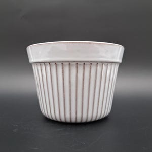 Puede incluir: Maceta de cerámica blanca con líneas verticales. La maceta tiene un fondo redondeado y un borde ancho.