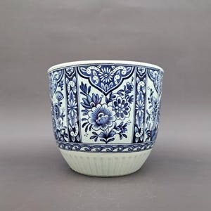 Może przedstawiać: Okrągła, biało-niebieska ceramiczna donica z kwiatowym wzorem. Donica ma powtarzający się wzór niebieskich kwiatów i liści na białym tle. Spód donicy ma pionowe żłobienia.