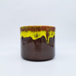 Pode incluir: Jardineira cilíndrica de cerâmica com acabamento brilhante. A borda superior apresenta uma faixa de esmalte amarelo que escorre sobre uma base marrom escura. A jardineira é projetada para uso interno ou externo.